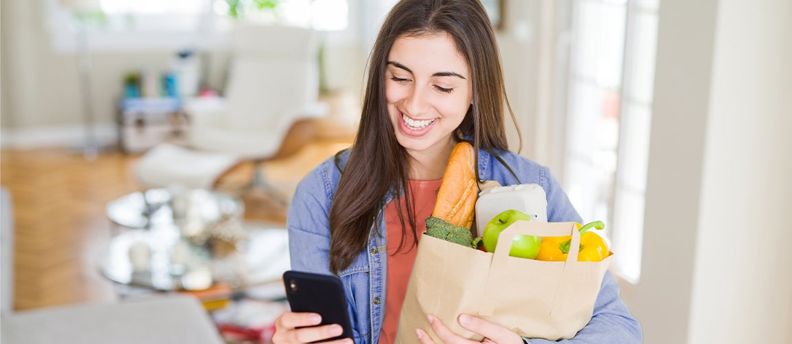 Young adult shopping for groceries while checking her bank account balance on her phone’s mobile banking app.