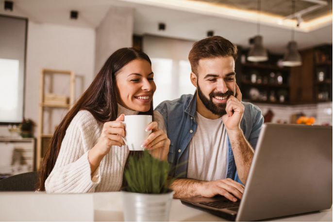 A couple sits at the kitchen table smiling while looking at a laptop screen